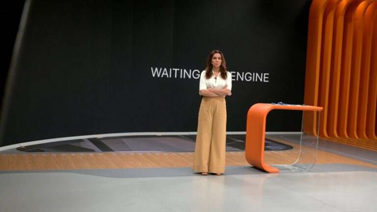 Apresentadora fica de braços cruzados ao lado de bancada laranja com tela exibindo mensagem waiting engine no estúdio do Bom Dia Brasil
