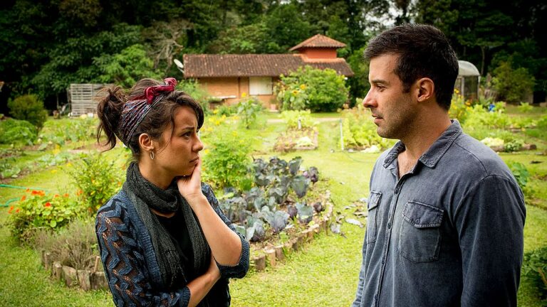 Mulher e homem conversam em horta ao ar livre com casa ao fundo em cena de novela