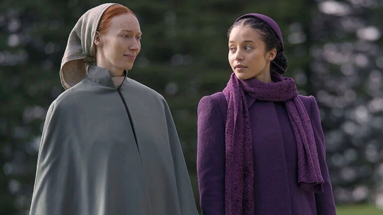 Duas mulheres caminham lado a lado e trocam olhares em cena de série ao ar livre com árvores ao fundo