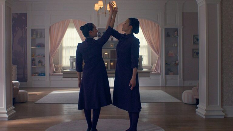 Duas jovens de vestidos escuros levantam as mãos juntas em sala elegante com cortinas claras ao fundo