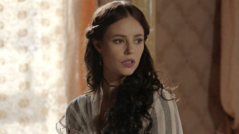 Mulher com cabelo ondulado preso de lado olha para trás com expressão tensa em cena de novela ambientada em casa