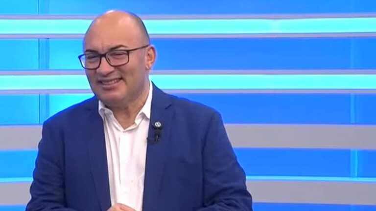 Homem de terno e óculos sorri durante participação em programa de TV com cenário azul iluminado