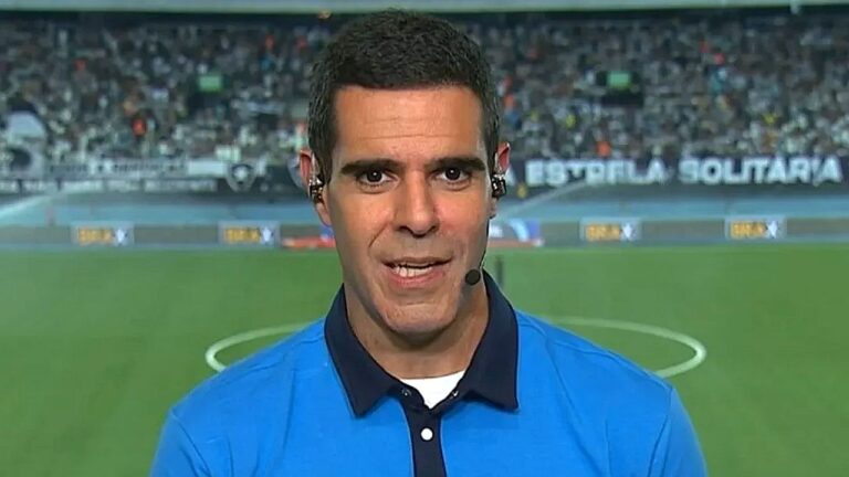 Repórter esportivo com microfone e ponto eletrônico fala diante de campo de futebol com arquibancada ao fundo