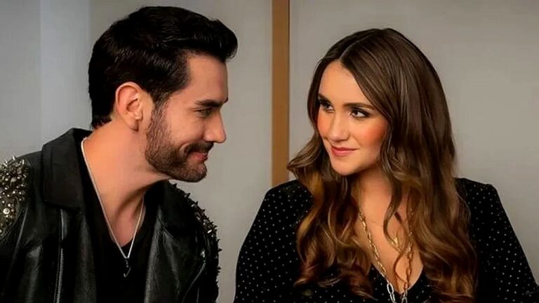 Texto Alternativo disse: Homem e mulher trocam olhares e sorriem sentados lado a lado em cena de série ou novela