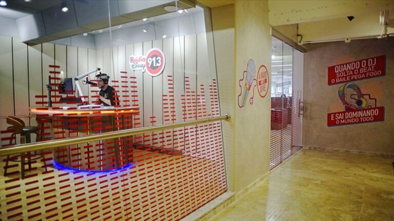Estúdio da Rádio Disney 91.3 com locutor ao microfone em cabine envidraçada e corredor ao lado