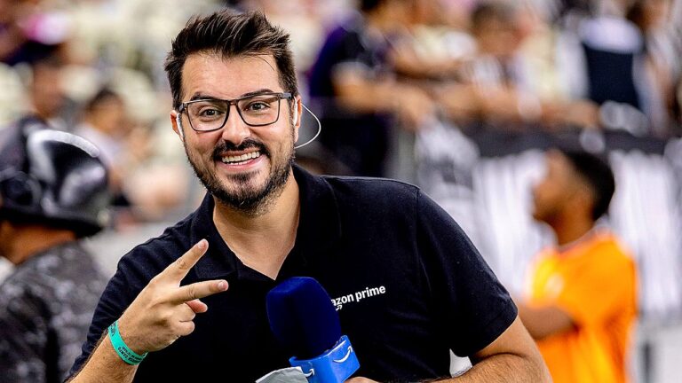 Repórter sorri e faz gesto de paz segurando microfone em cobertura de evento com público ao fundo