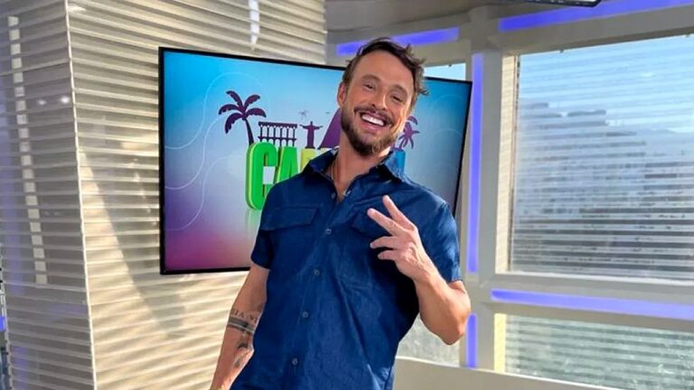 Apresentador sorri e faz gesto de paz em estúdio com tela do programa exibindo paisagem do Rio de Janeiro