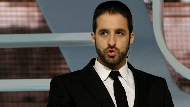 Apresentador de terno e gravata fala no palco de programa de TV com microfone preso ao rosto