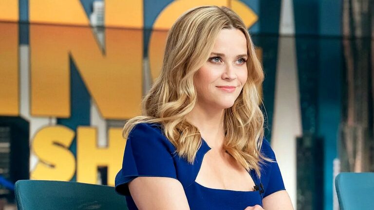 Atriz Reese Witherspoon sorri durante participação em programa de TV em estúdio com cenário colorido ao fundo