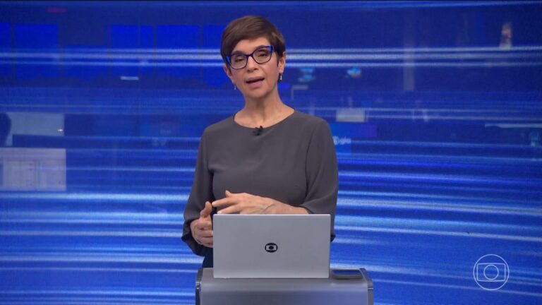 Renata Lo Prete, com óculos, fala ao lado de notebook no estúdio do Jornal da Globo, com telão azul ao fundo.