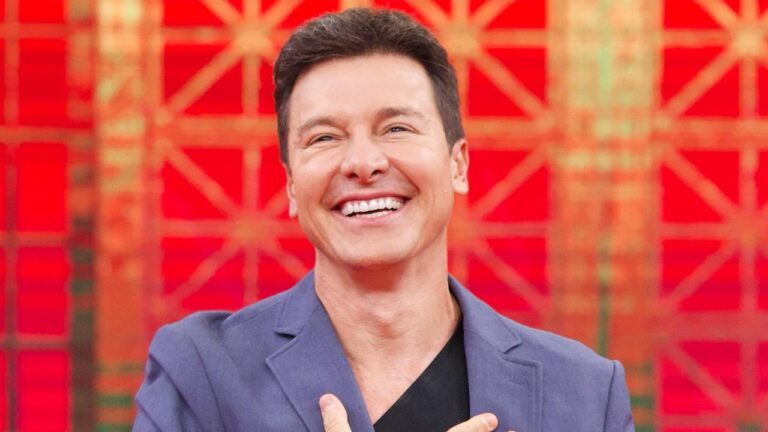 Rodrigo Faro sorrindo em estúdio com fundo vermelho geométrico durante gravação de programa de televisão