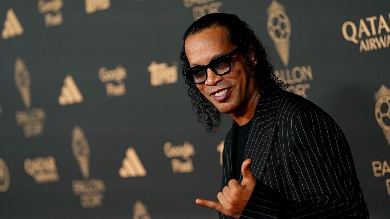 Ronaldinho Gaúcho posa sorrindo com gesto de mão em evento da Bola de Ouro com logos ao fundo