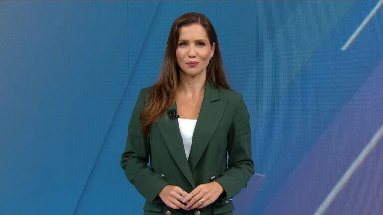 Fabíola Corrêa, apresentadora de telejornal com blazer verde, fala no estúdio do SBT Notícias durante transmissão ao vivo, com fundo azul minimalista como cenário.
