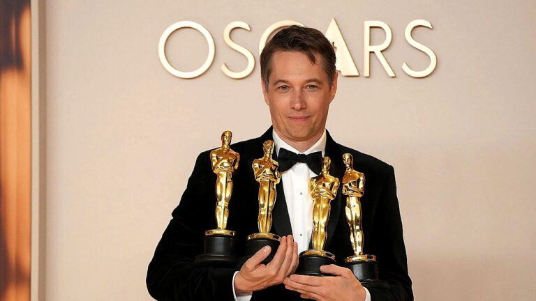 Homem de terno segura quatro estatuetas do Oscar durante premiação com painel escrito Oscars ao fundo