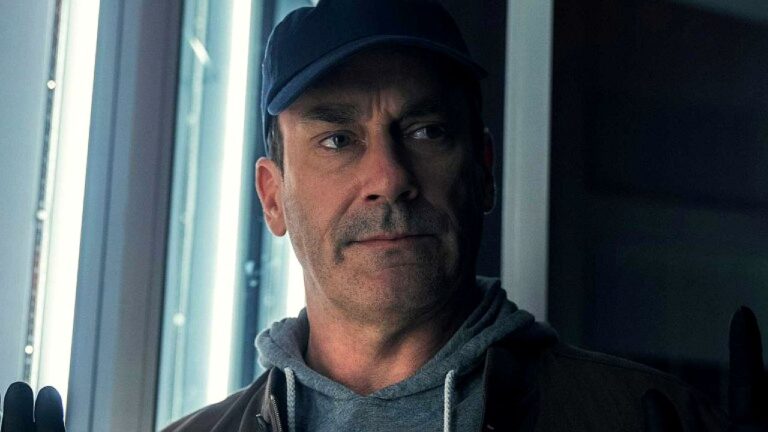 Homem de boné e moletom observa pela janela com expressão séria em cena de série dramática