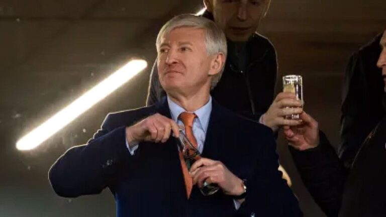 Homem de terno e gravata abre garrafa enquanto outro segura copo em cena com iluminação de estúdio ao fundo