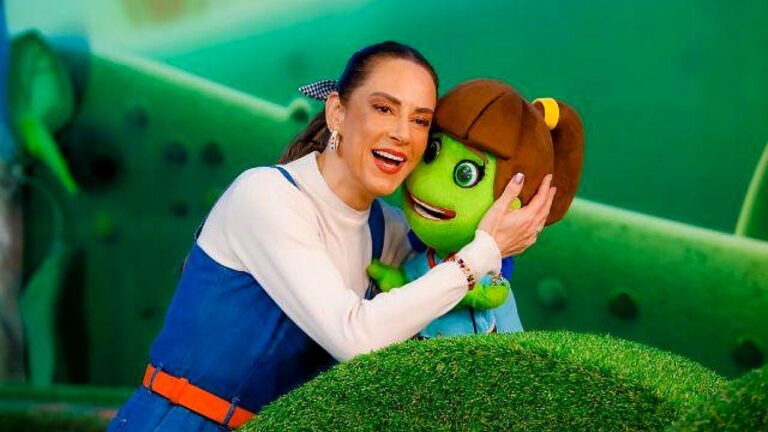 Apresentadora abraça personagem infantil verde com laço no cabelo em cenário colorido de programa de TV
