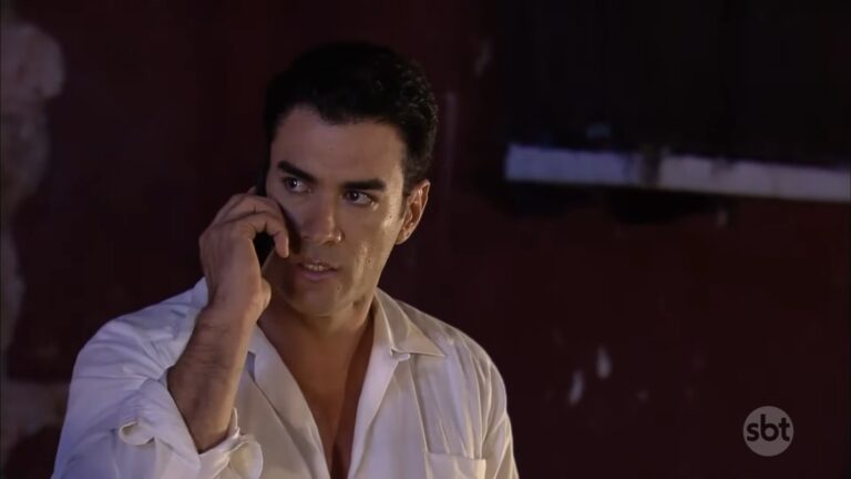 Homem fala ao celular com expressão tensa em cena de Sortilégio, novela do SBT, ambientada em local fechado.