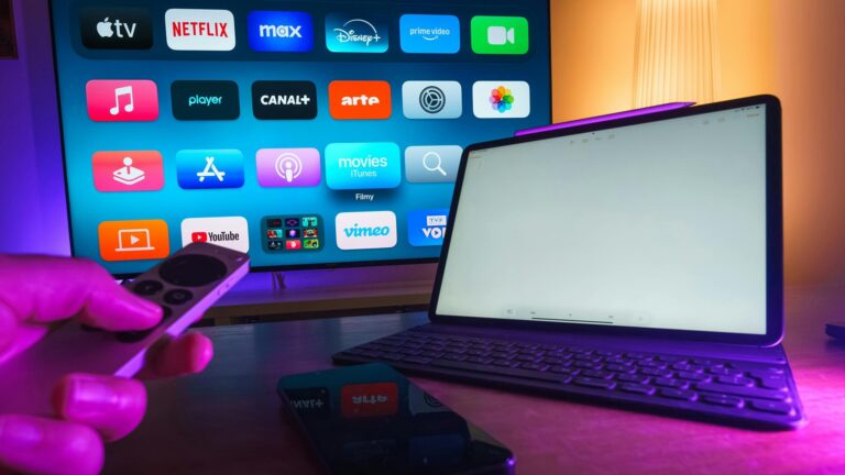 Tela de smart TV com apps Netflix Disney Plus e YouTube enquanto pessoa segura controle remoto diante de tablet sobre mesa