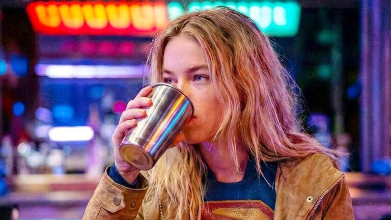 Mulher loira bebe em copo metálico em bar iluminado com luzes coloridas ao fundo em cena de série ou filme