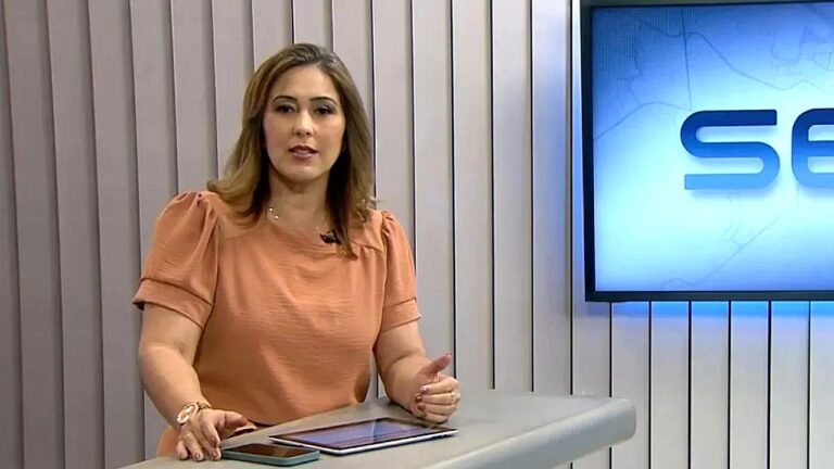 Apresentadora de blusa laranja fala em bancada de telejornal com tablet sobre a mesa e telão ao lado