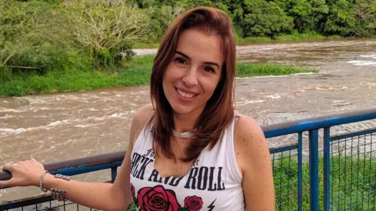 Suzane von Richthofen sorri apoiada em grade com rio e vegetação ao fundo usando camiseta clara sem mangas