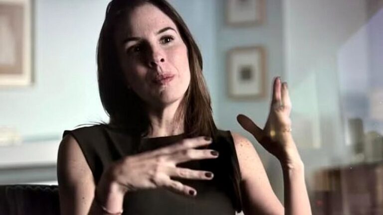 Mulher de blusa preta gesticula durante entrevista em ambiente interno com quadros ao fundo