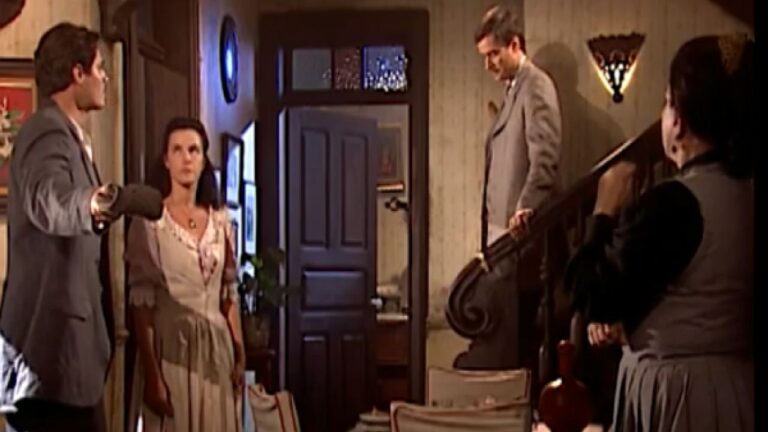 Homem aponta arma para mulher enquanto outros observam em sala de casa em cena tensa de novela