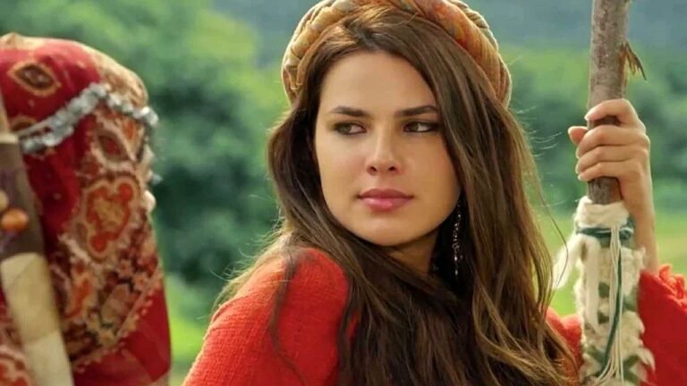 Mulher com traje típico e lenço na cabeça segura cajado em cenário rural com expressão desconfiada