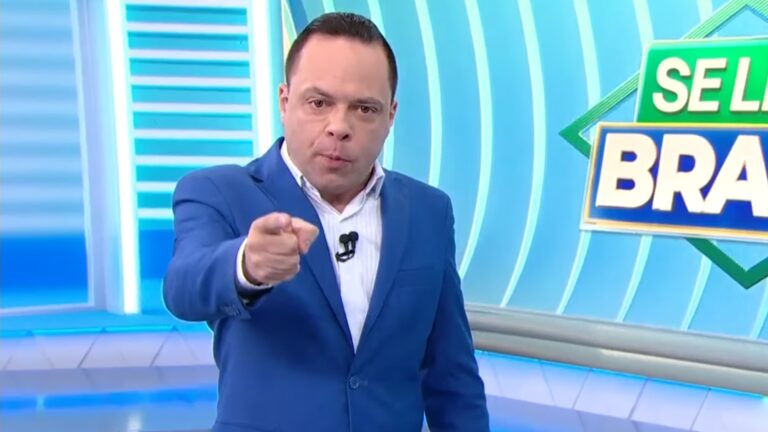 Apresentador Thiago Gardinali, vestindo um terno azul, aponta para a câmera enquanto apresenta o jornal Se Liga, Brasil, em cenário moderno, no SBT.