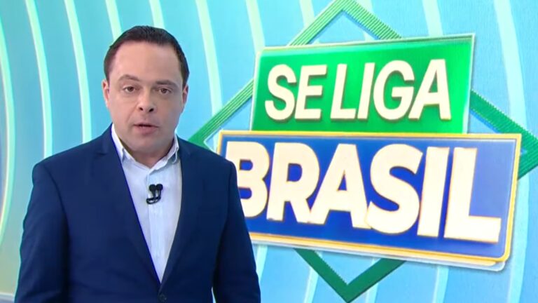 Thiago Gardinali, apresentador do Se Liga, Brasil, veste um terno azul e fala no estúdio do SBT com logotipo do programa ao fundo.