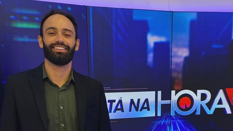 Apresentador sorri em estúdio diante de telão com o logo do programa Tá Na Hora ao fundo