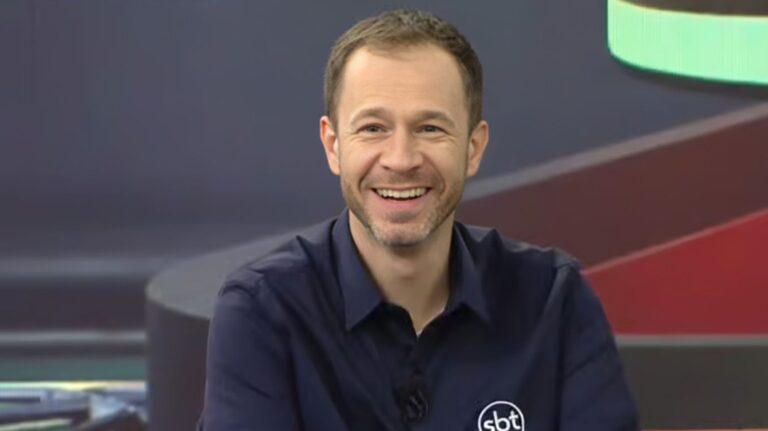 Tiago Leifert, apresentador do SBT, sorri durante participação em programa de TV em estúdio com cenário colorido ao fundo.