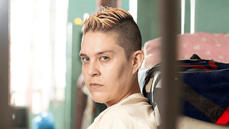 Pessoa de cabelo curto raspado olha para trás com expressão séria em ambiente interno de cena dramática