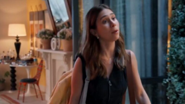 Mulher com mochila fala na porta de casa em cena de novela com sala iluminada ao fundo