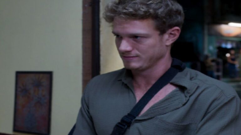 Homem com tipoia no braço observa alguém com expressão tensa em cena de novela ambientada em interior de casa