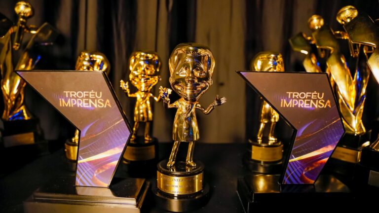 Estatuetas douradas do Troféu Imprensa dispostas sobre mesa com placas do prêmio e fundo com cortinas em evento de premiação
