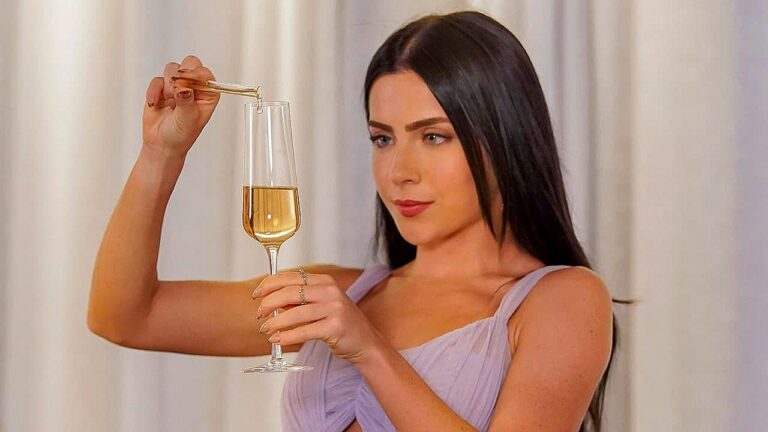 Mulher segura taça com líquido dourado enquanto usa conta-gotas em cena com foco em experimento ou bebida