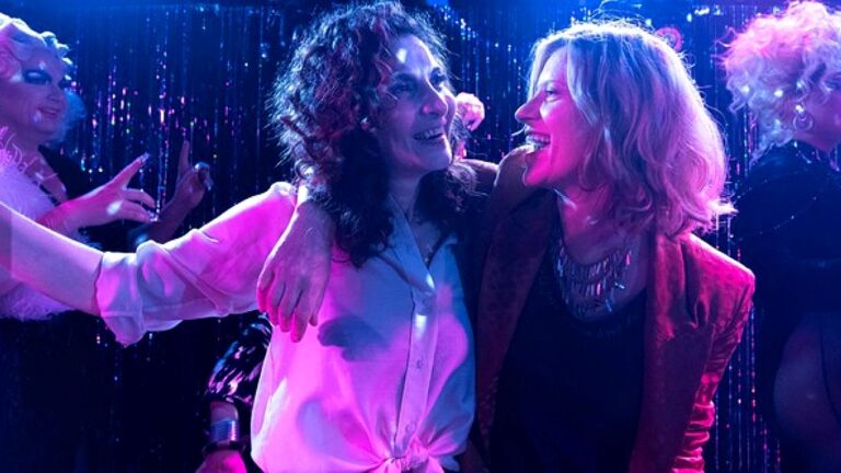 Duas mulheres sorrindo abraçadas em pista de dança com luzes coloridas e outras pessoas ao fundo em cena de série