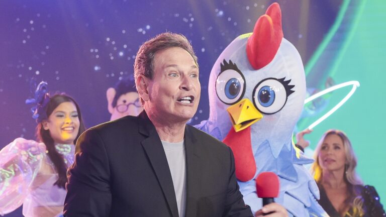 Luís Ricardo, apresentador do Viva a Noite, usa microfone vermelho ao lado de mascote de galinha em palco com dançarinas ao fundo.