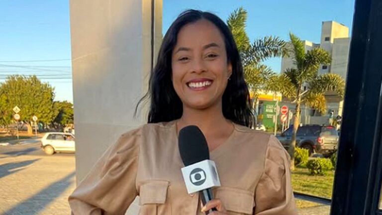 Repórter sorri segurando microfone da TV Globo durante entrada ao vivo em rua com carros e prédios ao fundo