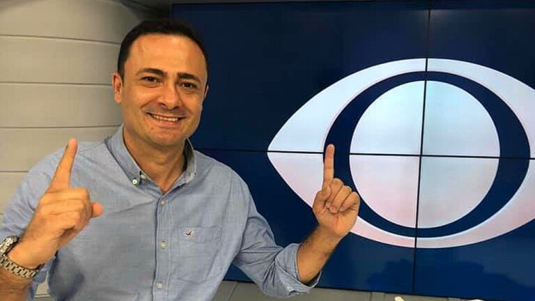 Apresentador sorri e aponta para logotipo da Band exibido em tela no estúdio de TV