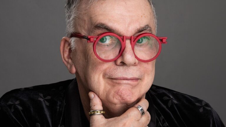 Walcyr Carrasco posa com óculos redondos vermelhos apoiando o queixo na mão em retrato de estúdio com fundo neutro