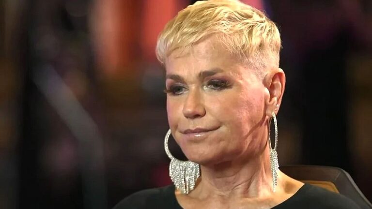 Apresentadora de cabelo curto e loiro com expressão séria em programa de TV com iluminação de estúdio