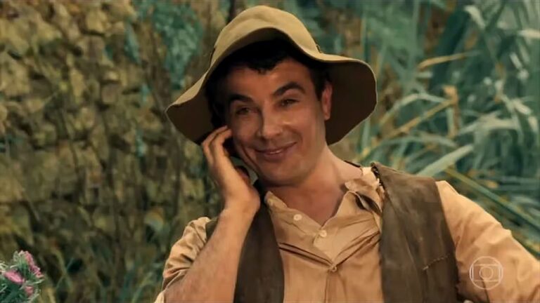 Homem com chapéu de palha sorri enquanto fala ao telefone em cenário rural em cena de novela