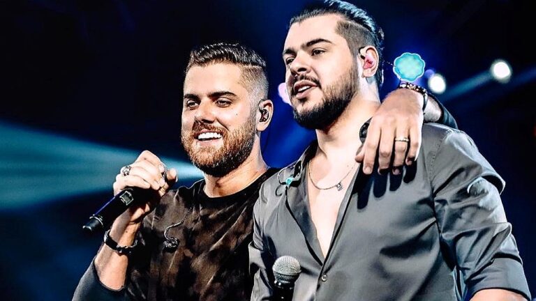 Dupla sertaneja canta abraçada no palco com microfones durante show ao vivo com luzes ao fundo