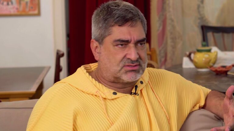 Homem de moletom amarelo fala com expressão séria sentado em sala de estar com mesa e objetos ao fundo