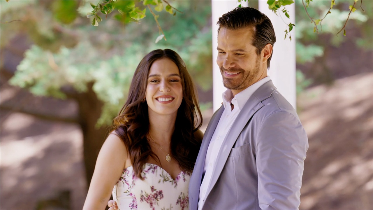 Casal sorri abraçado em cena romântica de A História de Joana, nova novela do SBT, ao ar livre com árvores e varanda ao fundo.
