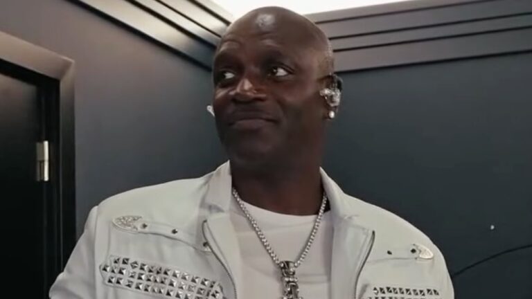 Akon usa jaqueta branca com aplicações metálicas e fones de ouvido durante aparição em bastidores de evento ou gravação