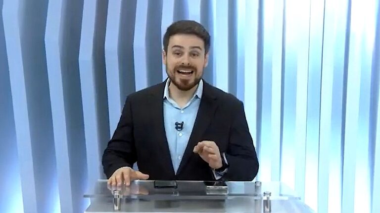 Apresentador fala atrás de bancada em estúdio com fundo azul durante programa de TV